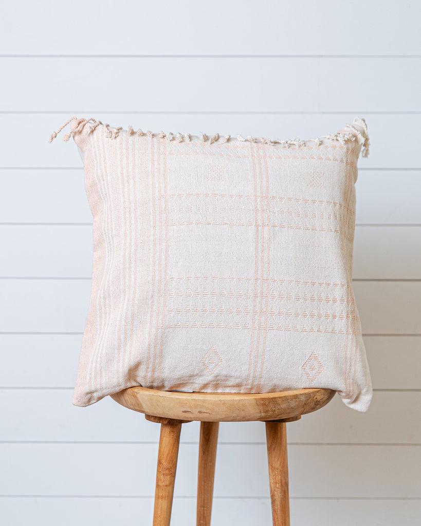 Ivory cotton cushion cover with soft blush-pink threads and fringed edges on wooden stool