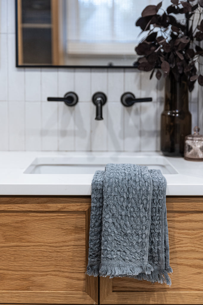 Slate blue waffle-knit cotton hand towel draped over white bathroom sink with wood cabinet