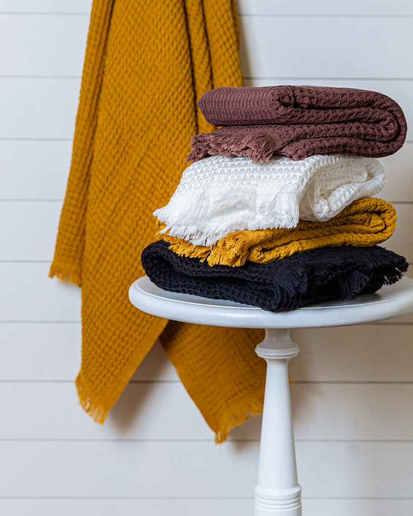 Stack of Turkish waffle bath towels in mustard, burgundy, white, and deep navy, folded on a stool